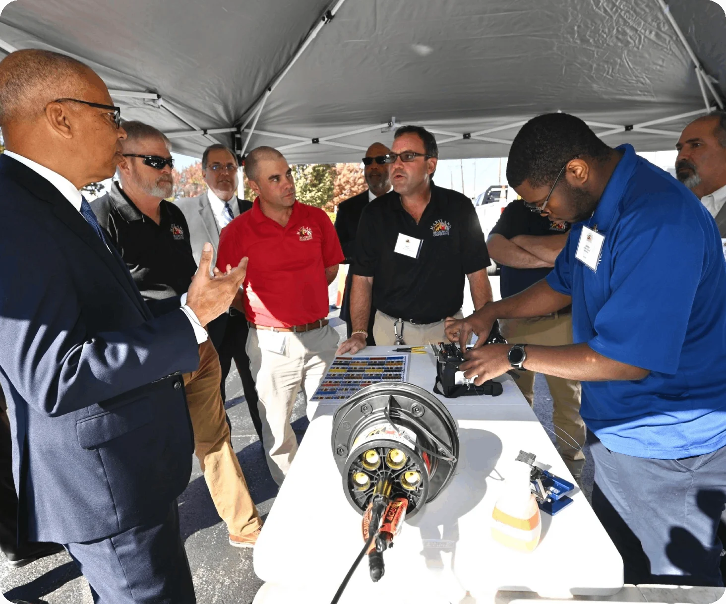 A group of men stand under a tent, intently watching a demonstration of fiber optic networking equipment on a table. One man in a blue shirt is actively working on the equipment while others observe and discuss.