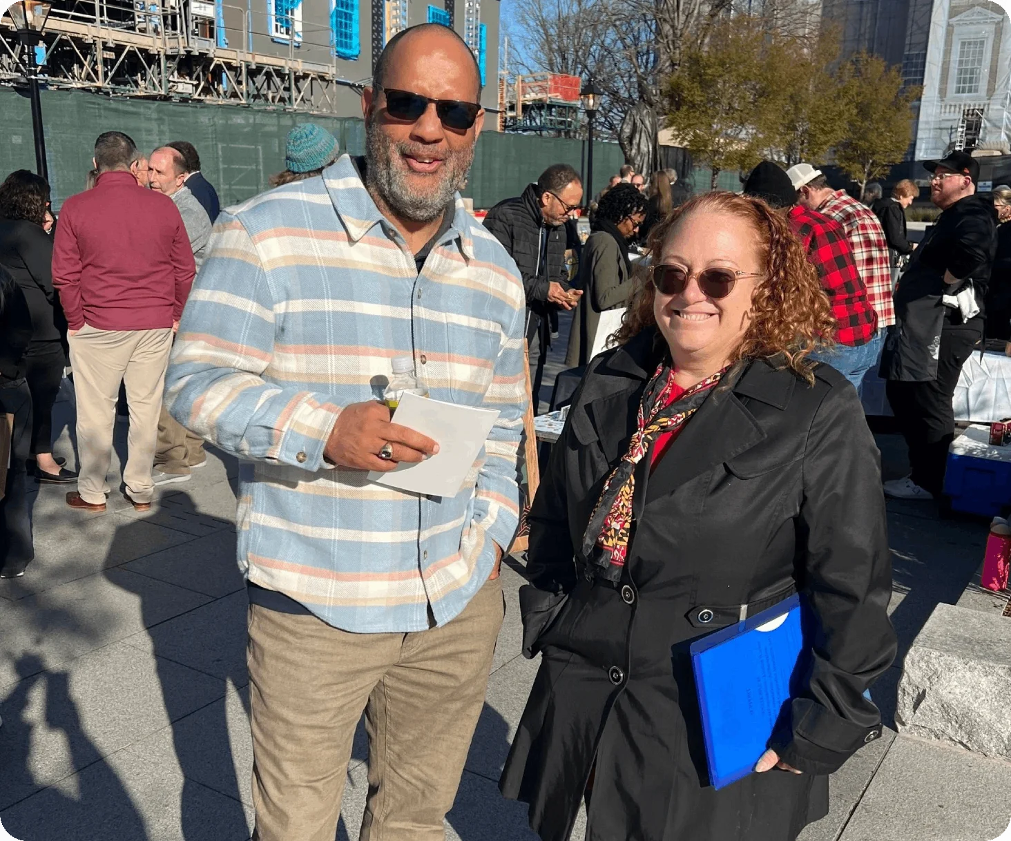 A man wearing sunglasses and holding a piece of paper stands next to a woman with curly hair holding a blue folder. They are outdoors in a crowd with some buildings in the background, possibly discussing fiber optic networking.