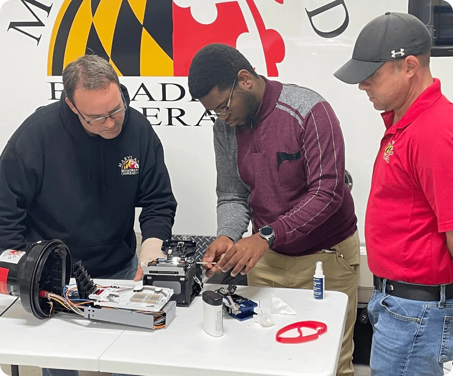 Three men are working on an electrical device related to fiber optic networking on a white table, with a partially visible logo on the wall in the background. One man observes, while the other two adjust the components carefully.