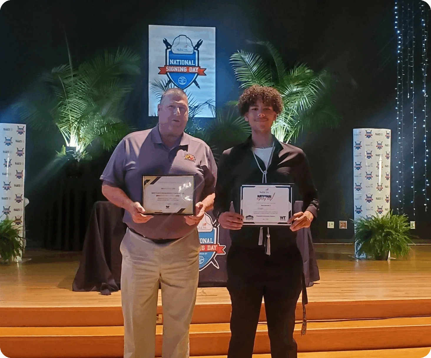 Two individuals stand on a stage holding certificates with a "National Signing Day" banner in the background, flanked by plants and decorative elements, highlighting their achievements in fiber optic networking.