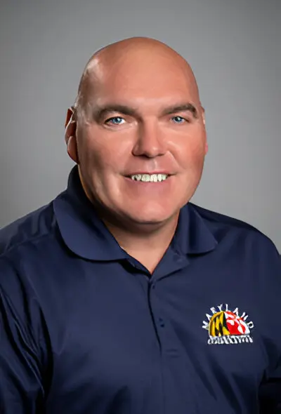 A man with a bald head, wearing a navy blue polo shirt featuring the Maryland Terrapins logo, smiles at the camera against a plain grey background, embodying the confident look of someone who just secured reliable broadband access.