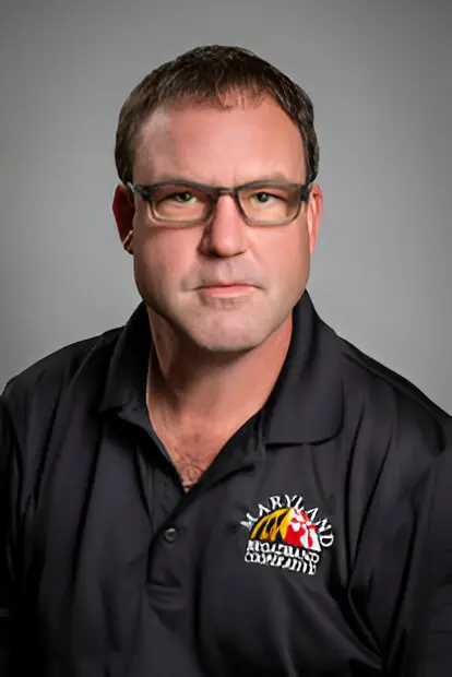 A man with glasses is wearing a black polo shirt with a Maryland-themed logo. He is looking directly at the camera against a plain gray background, symbolizing the clarity and focus needed for widespread broadband access.