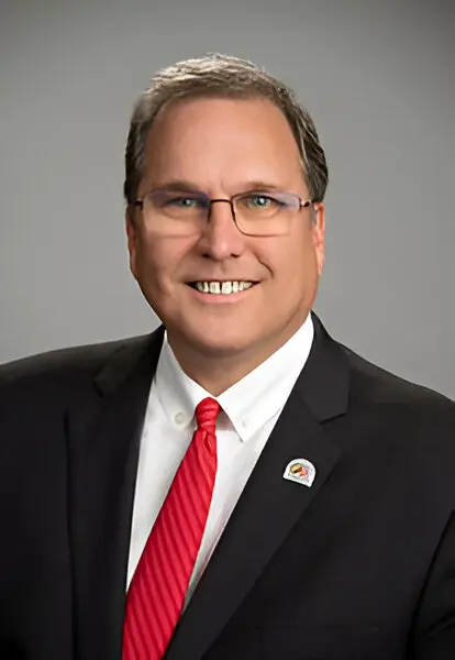 A man wearing a black suit, white shirt, and red tie, with glasses, smiles at the camera against a grey background, embodying the professionalism vital for efficient broadband access.