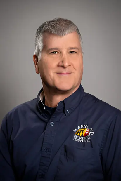 A person with short gray hair smiles at the camera, wearing a dark blue shirt with a logo featuring a bee and text that reads "Larry's Broadband Access.