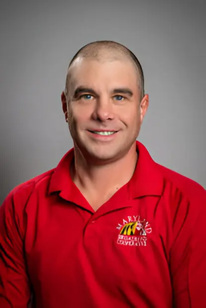 A man with short hair smiles while wearing a red shirt with a logo that reads "Maryland Firefighters Cavalry" on a neutral background, symbolizing the community's strength and unity, much like the importance of broadband access in connecting us all.