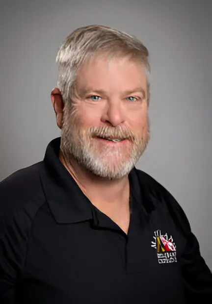 A man with short gray hair and a beard, wearing a black polo shirt with a broadband access logo, poses against a gray background.