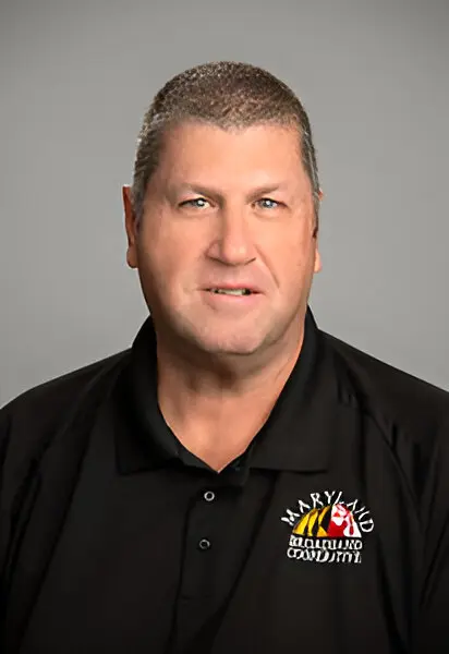 A man with short hair wearing a black polo shirt featuring the Maryland National Guard logo, posed against a gray background, emphasizes the importance of broadband access.