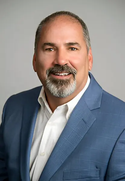 A man with short hair and a graying beard wears a blue suit jacket and white shirt. He is smiling and looking directly at the camera, exuding confidence reminiscent of a leader in broadband access. The background is plain gray.