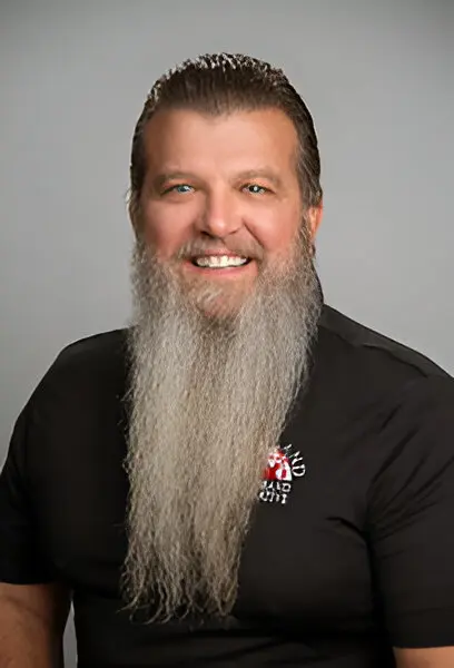 A man with a long grey beard, smiling, and wearing a black shirt with a broadband access logo on it, posed against a neutral background.