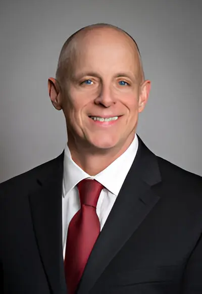 A man in a business suit with a red tie poses against a neutral background. He is smiling and bald, embodying the professional image of someone familiar with broadband access.