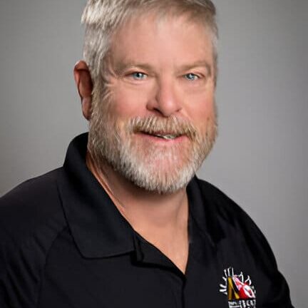 A man with short gray hair and a beard, wearing a black polo shirt with a broadband access logo, poses against a gray background.